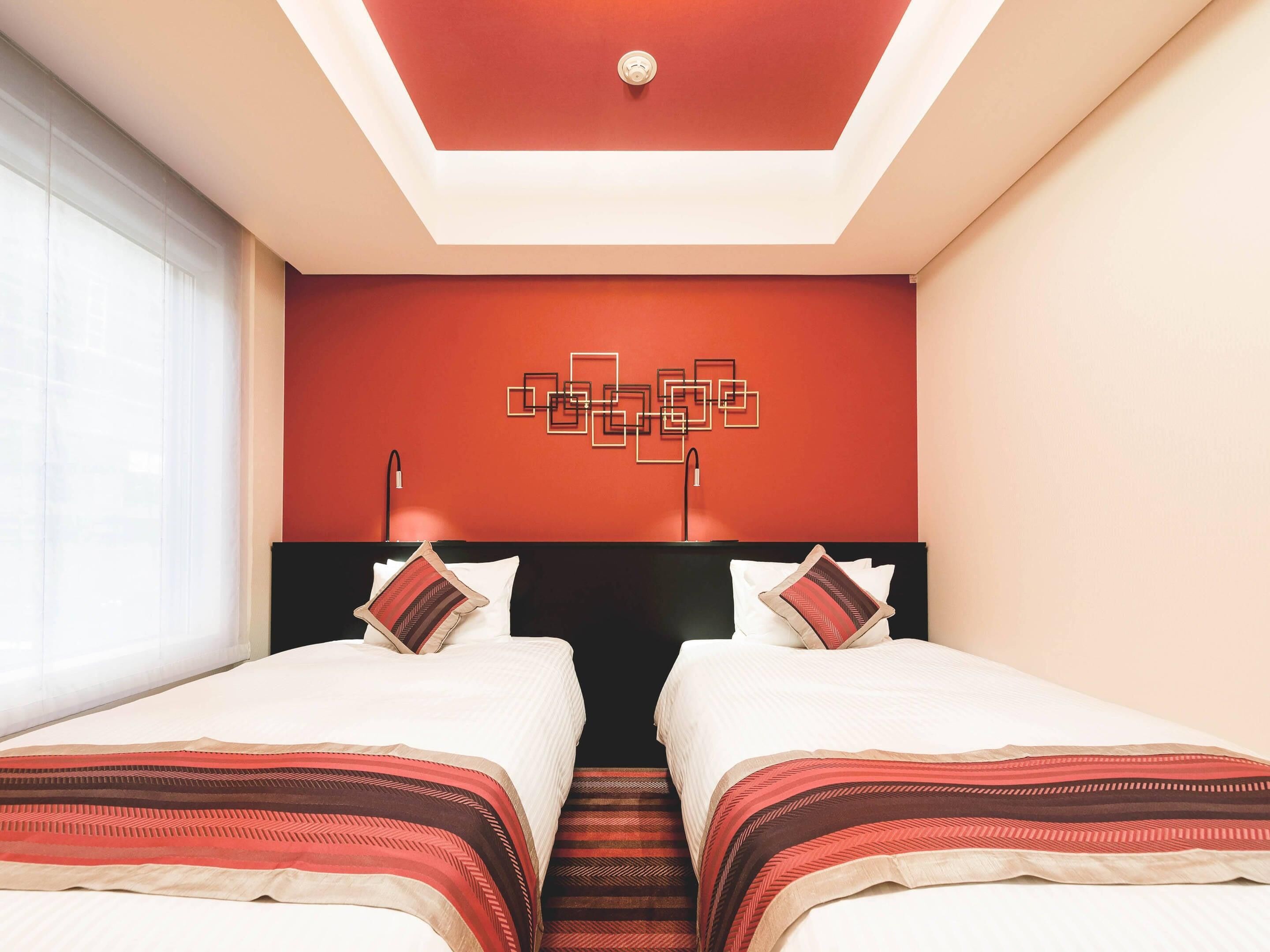 A hotel room with two beds, a red accent wall, a geometric wall art piece, and a ceiling light fixture.