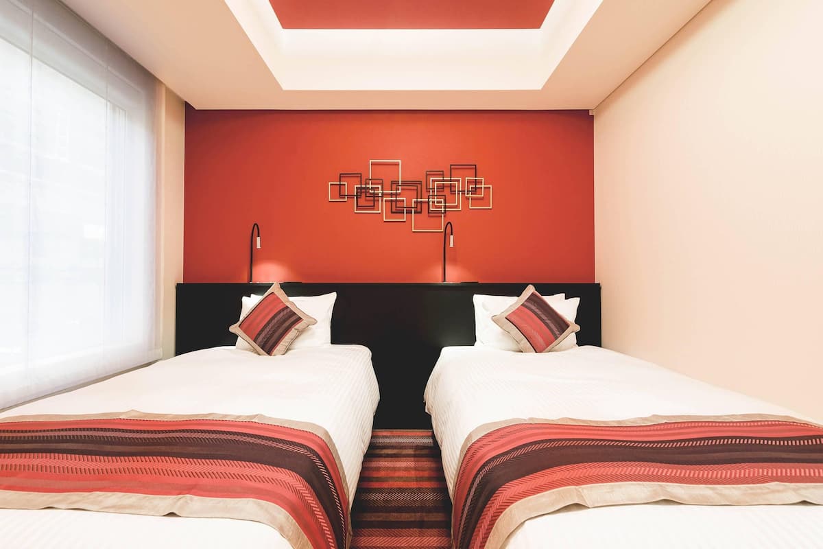 A hotel room with two beds, a red accent wall, a geometric wall art piece, and a ceiling light fixture.