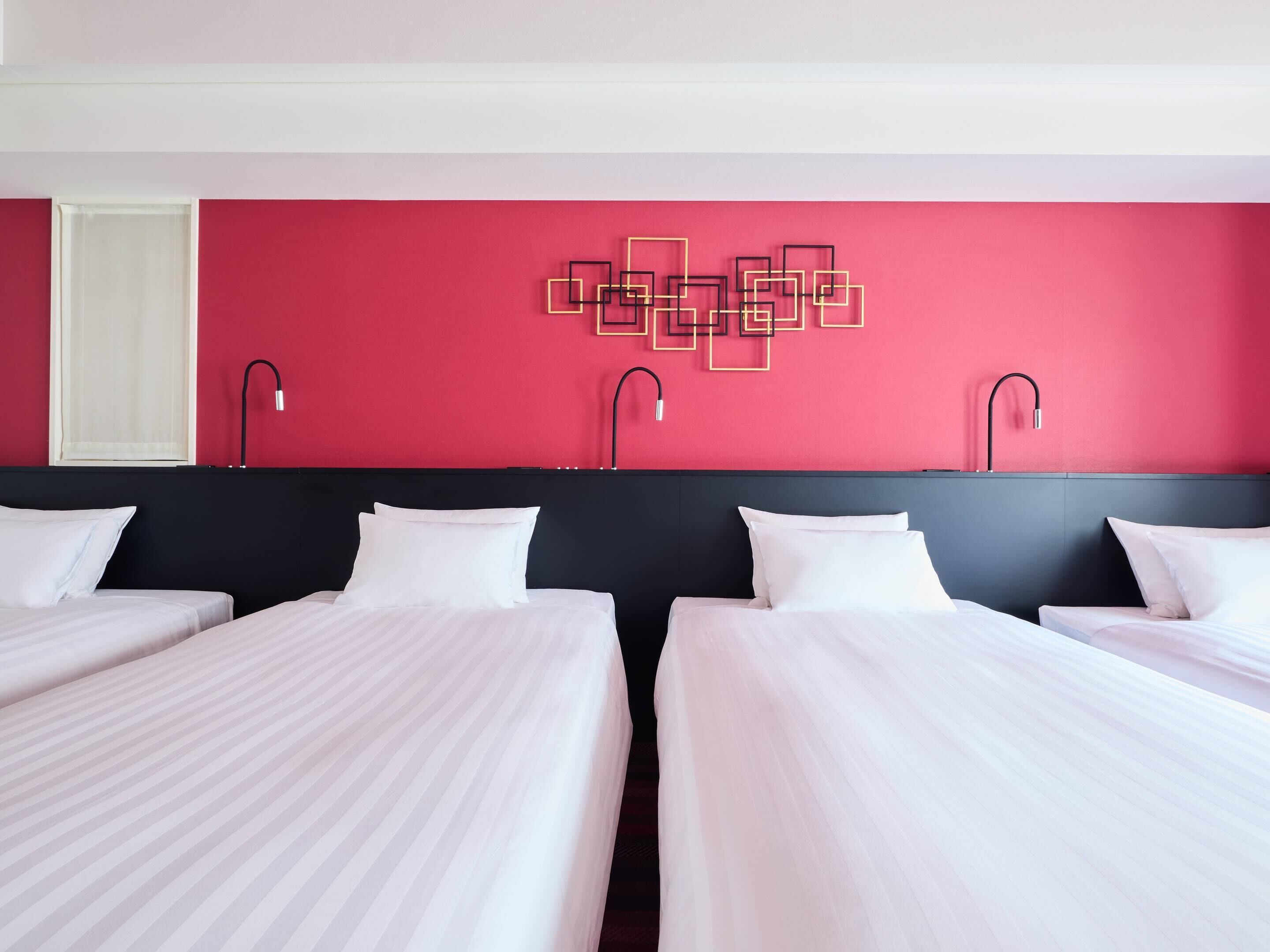 Habitación de hotel con dos camas, una pared de acento roja y una lámpara con diseño geométrico.