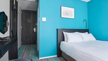 A hotel room with a bed, a desk with a telephone, a chair, a wall-mounted picture, and a door leading to another room.