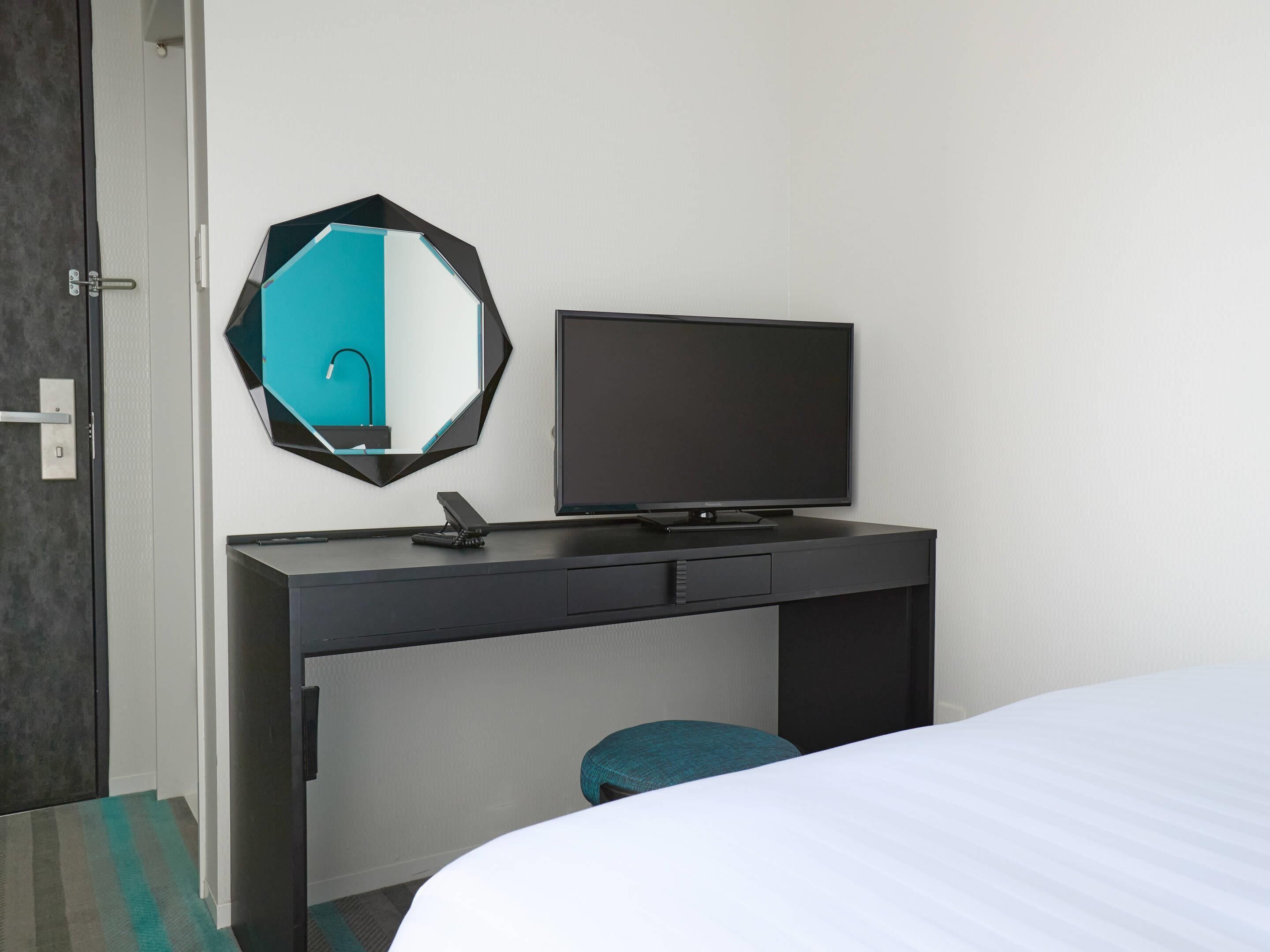 A hotel room with a bed, a desk with a flat-screen TV, a mirror, and a small stool.
