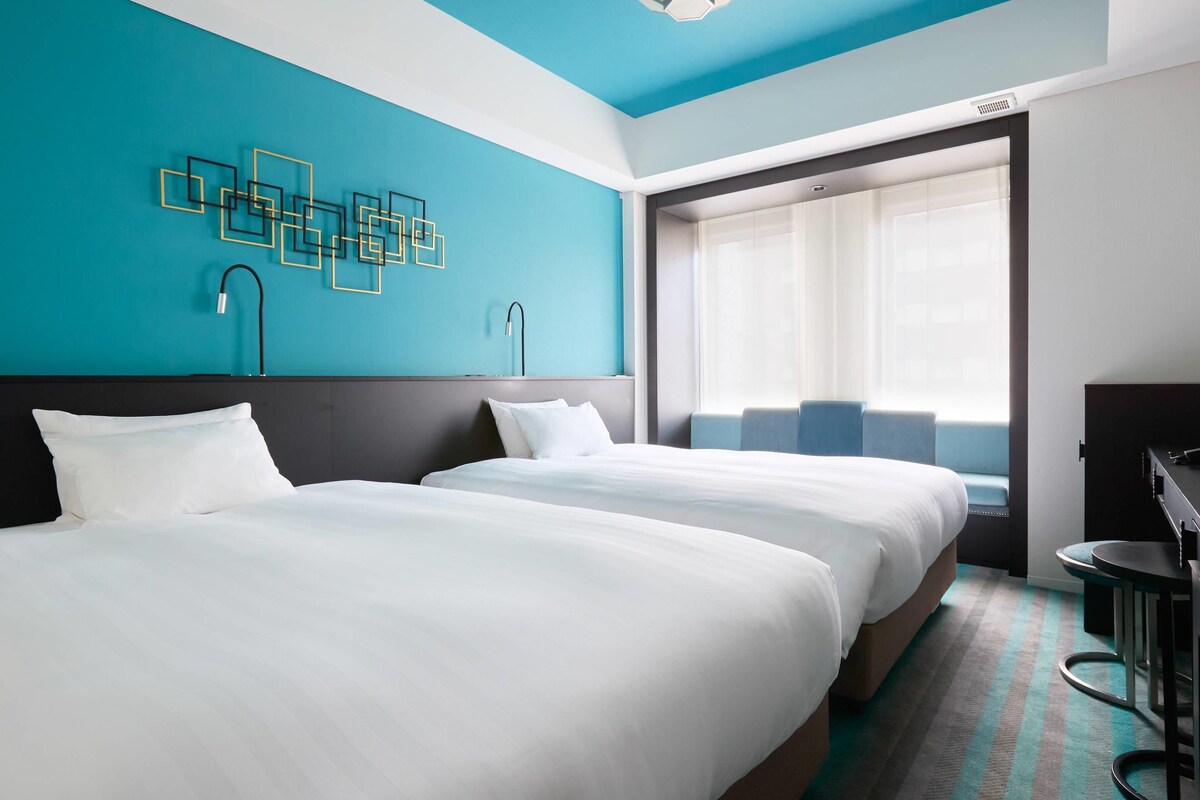 Une chambre d’hôtel avec deux lits, un bureau et une grande fenêtre avec des rideaux.