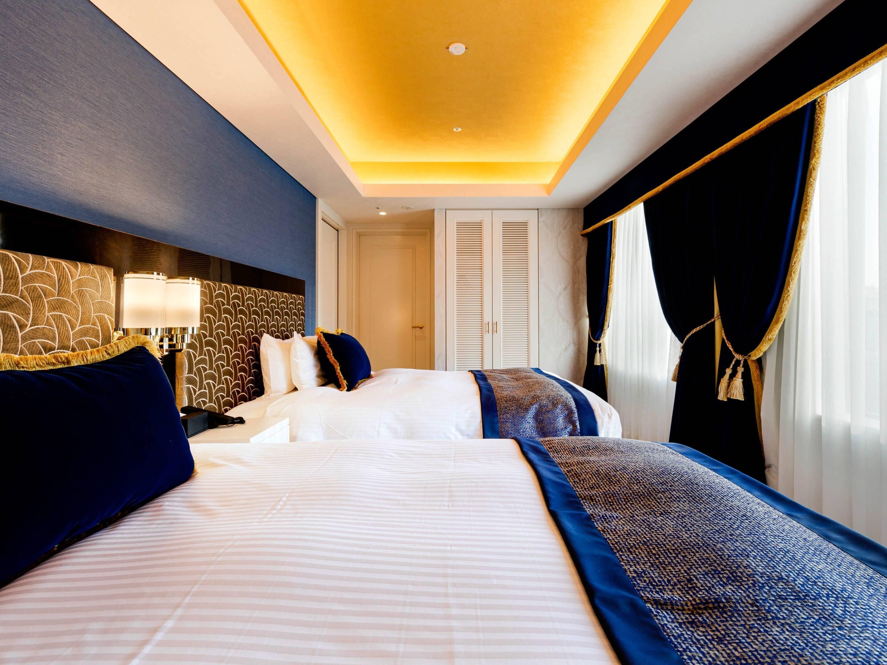 A hotel room with a large bed, a headboard with a geometric pattern, a bedside table with a lamp, and a view of the outside through a window with curtains.