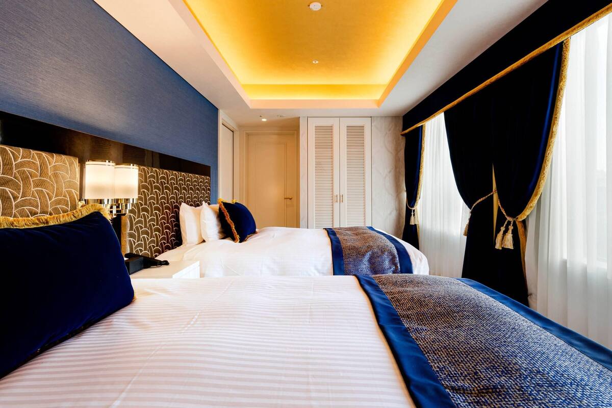 Une chambre d’hôtel avec un grand lit, une tête de lit ornée d’un motif géométrique, une table de chevet avec une lampe, et une vue sur l’extérieur par une fenêtre avec des rideaux.