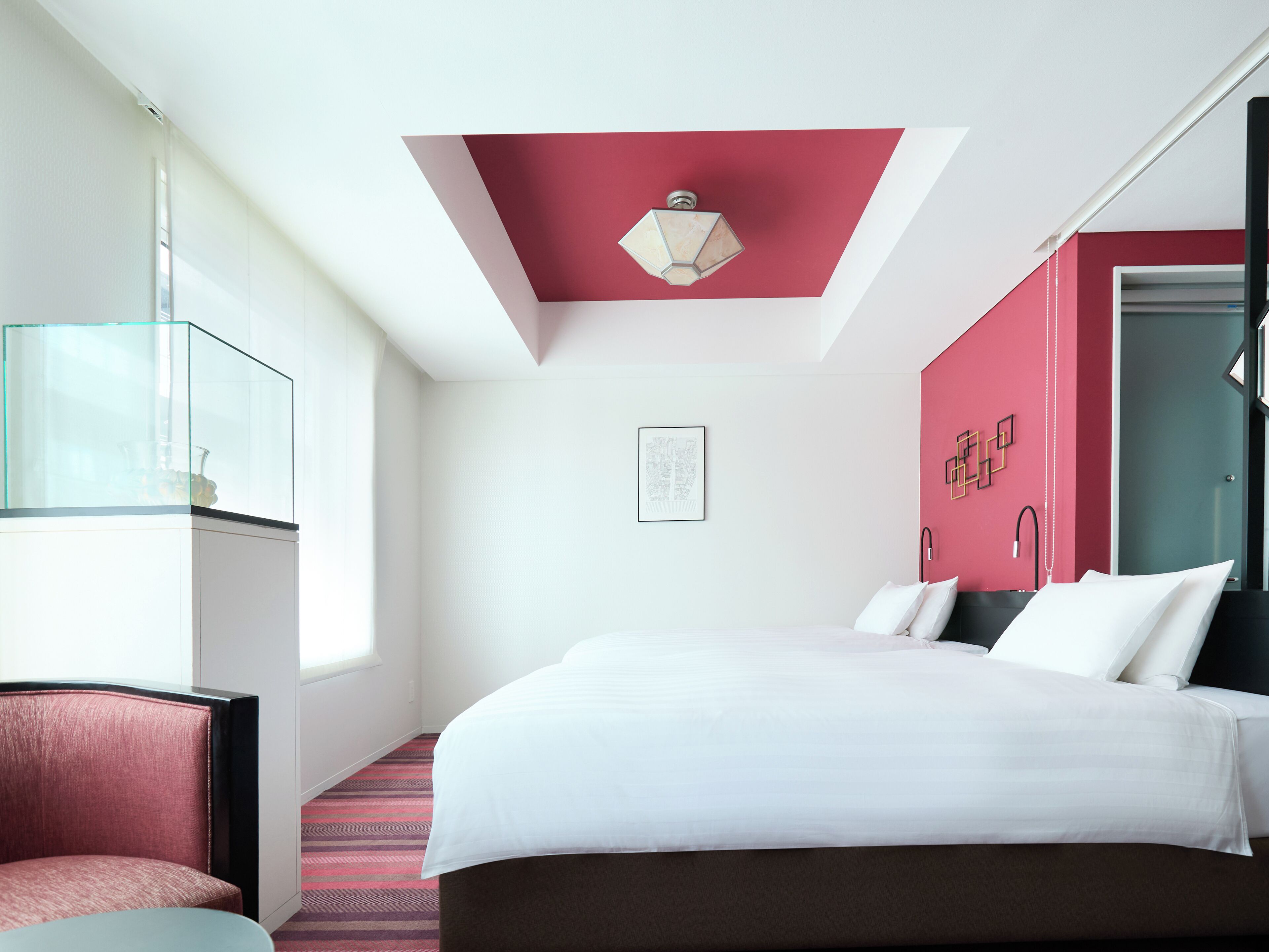 A hotel room with a bed, a chair, a glass partition, and a red accent wall.