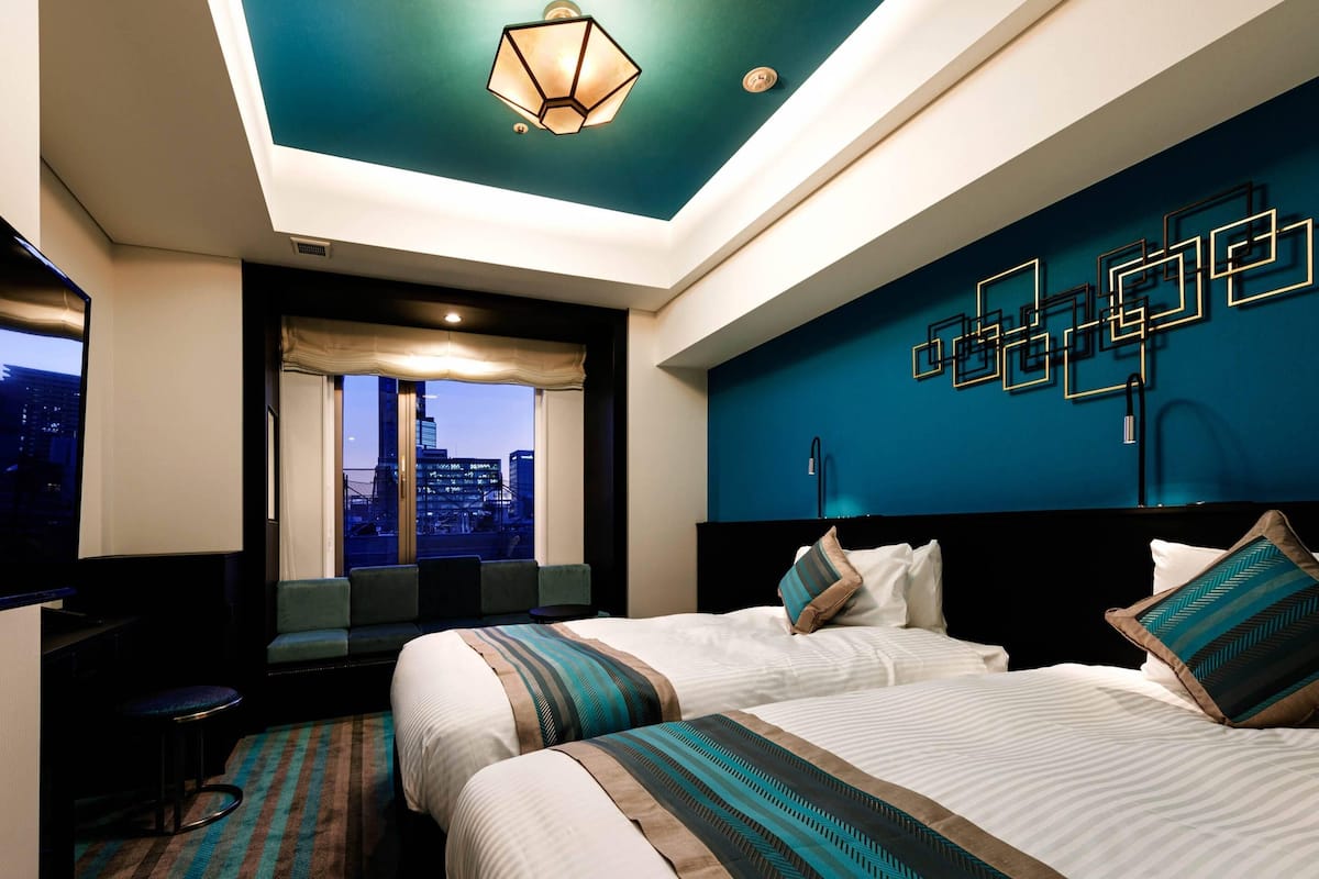 A hotel room with a city view, two beds, a desk, and a modern ceiling design.
