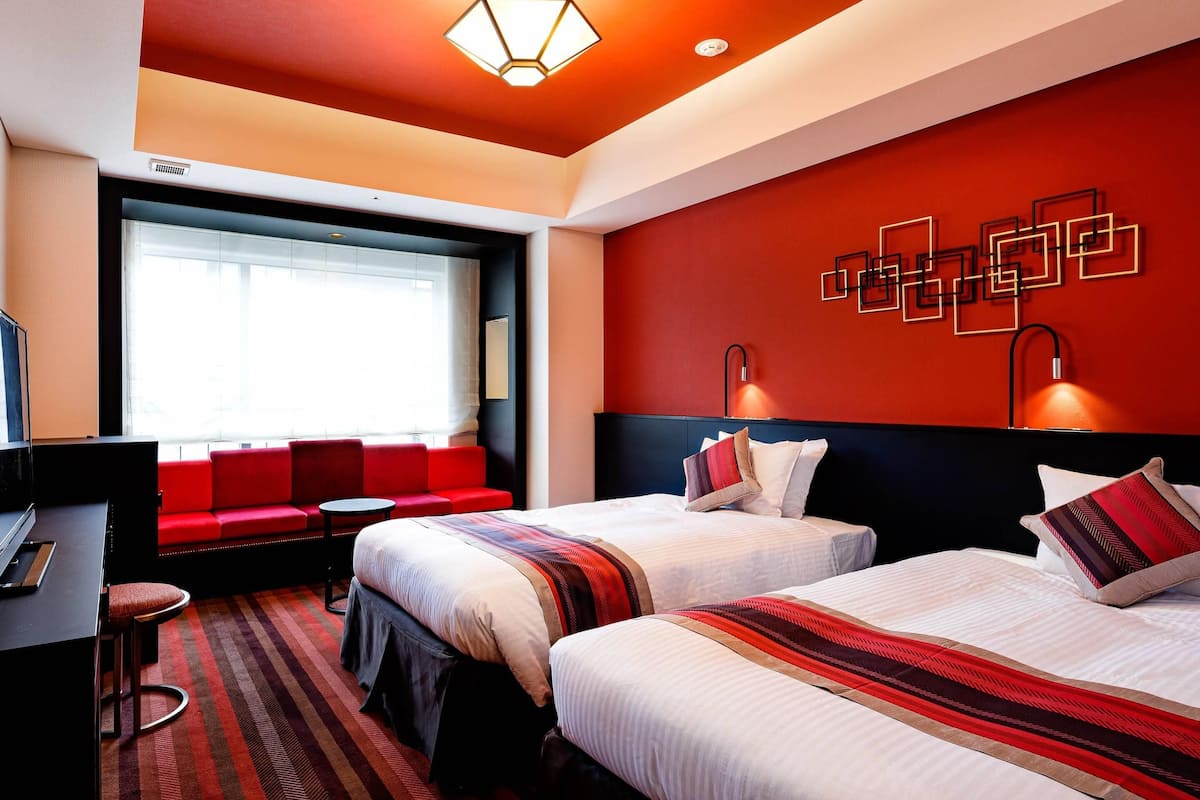 A hotel room with two beds, a desk, a TV, a sofa, and a large window.