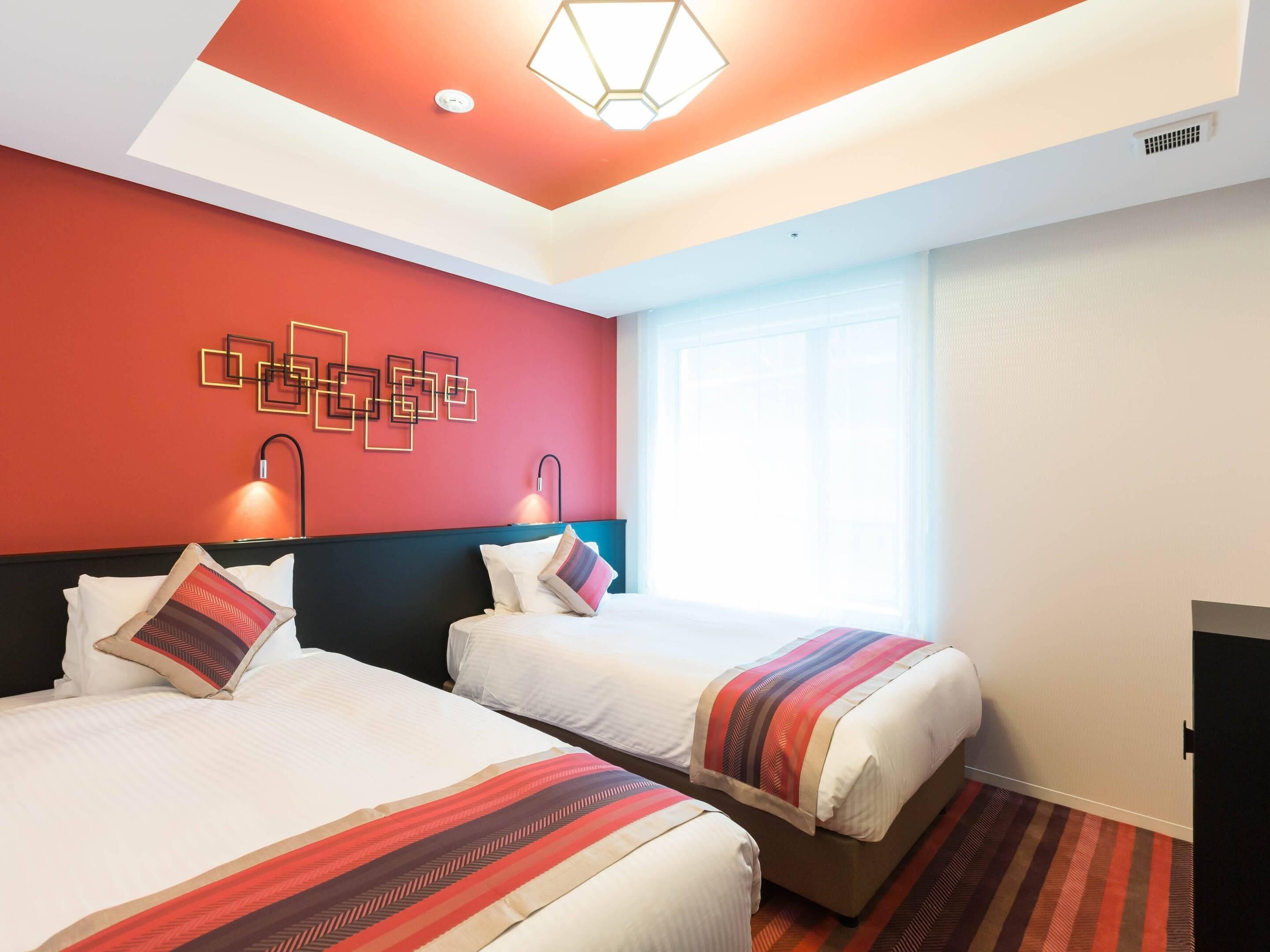 A hotel room with two beds, a red accent wall, a geometric light fixture, and a large window.
