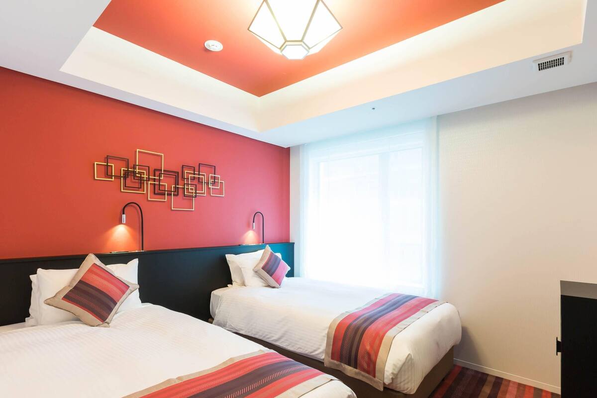 Une chambre d’hôtel avec deux lits, un mur d’accent rouge, un luminaire au design géométrique et une grande fenêtre.