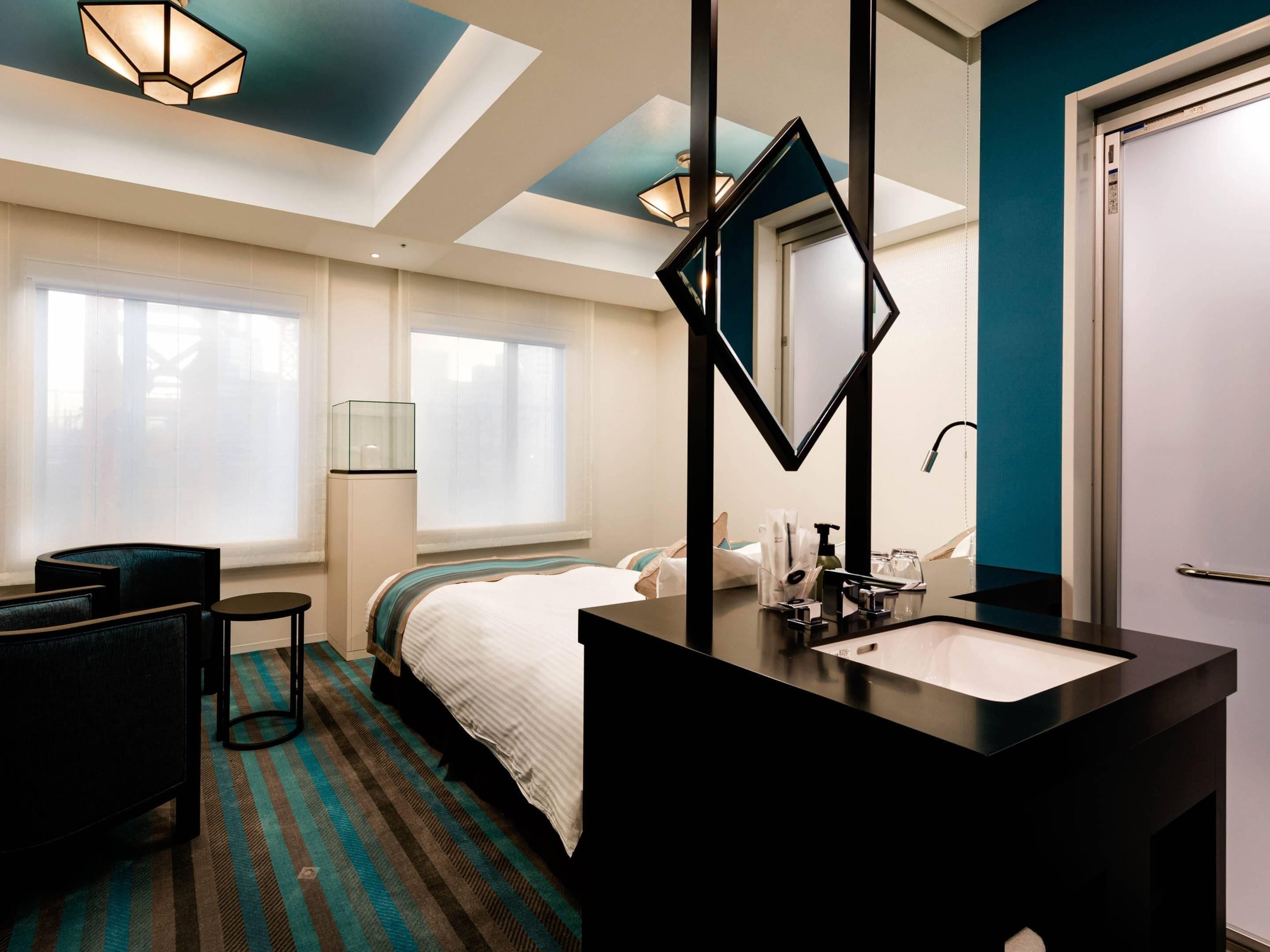 A hotel room with a bed, a black vanity with a mirror, a window with blinds, a chair, and a striped carpet.