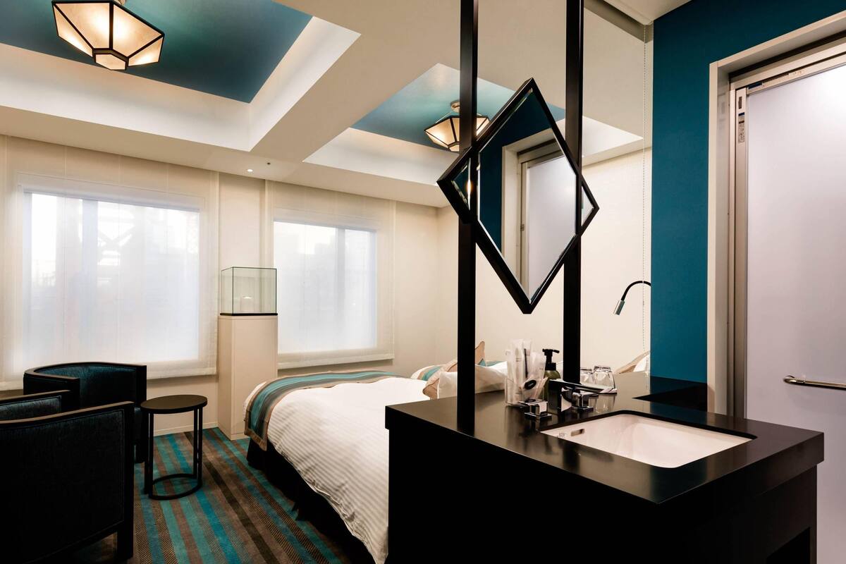 Une chambre d’hôtel avec un lit, une coiffeuse noire dotée d’un miroir, une fenêtre avec des stores, une chaise et une moquette à rayures.