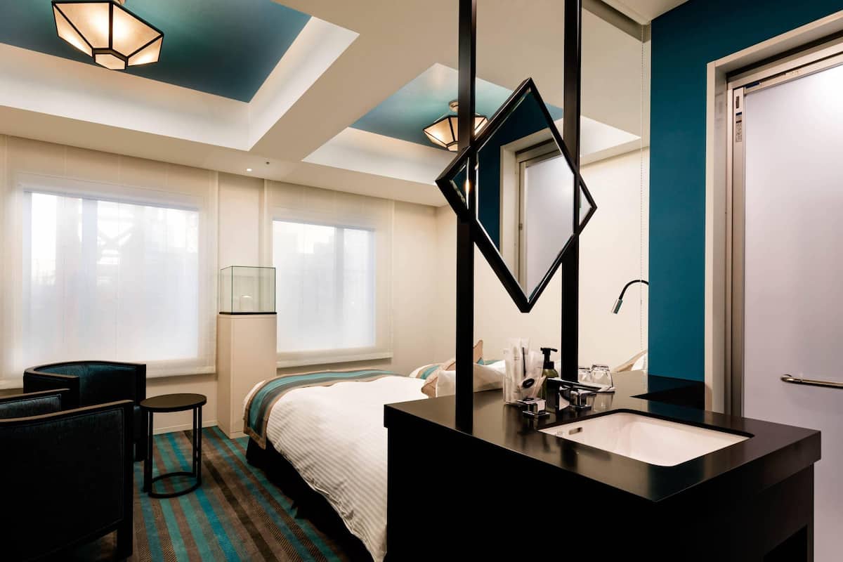 A hotel room with a bed, a black vanity with a mirror, a window with blinds, a chair, and a striped carpet.