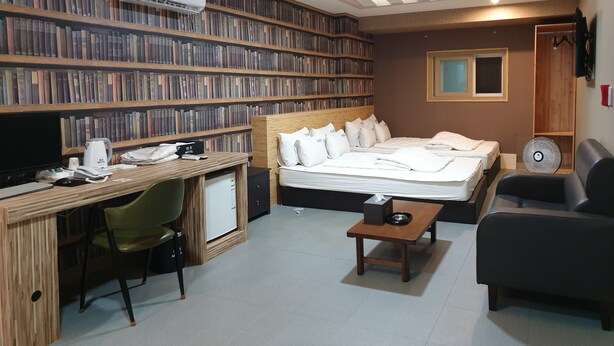 A hotel room with a large bookshelf, a bed with white bedding, a desk with a computer, and a small table.