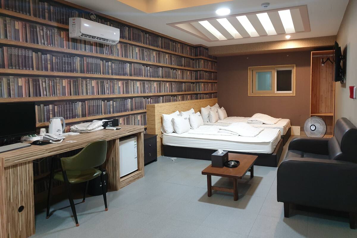 Quarto de hotel com uma grande estante de livros, uma cama com roupa de cama branca, uma escrivaninha com um computador e uma pequena mesa.