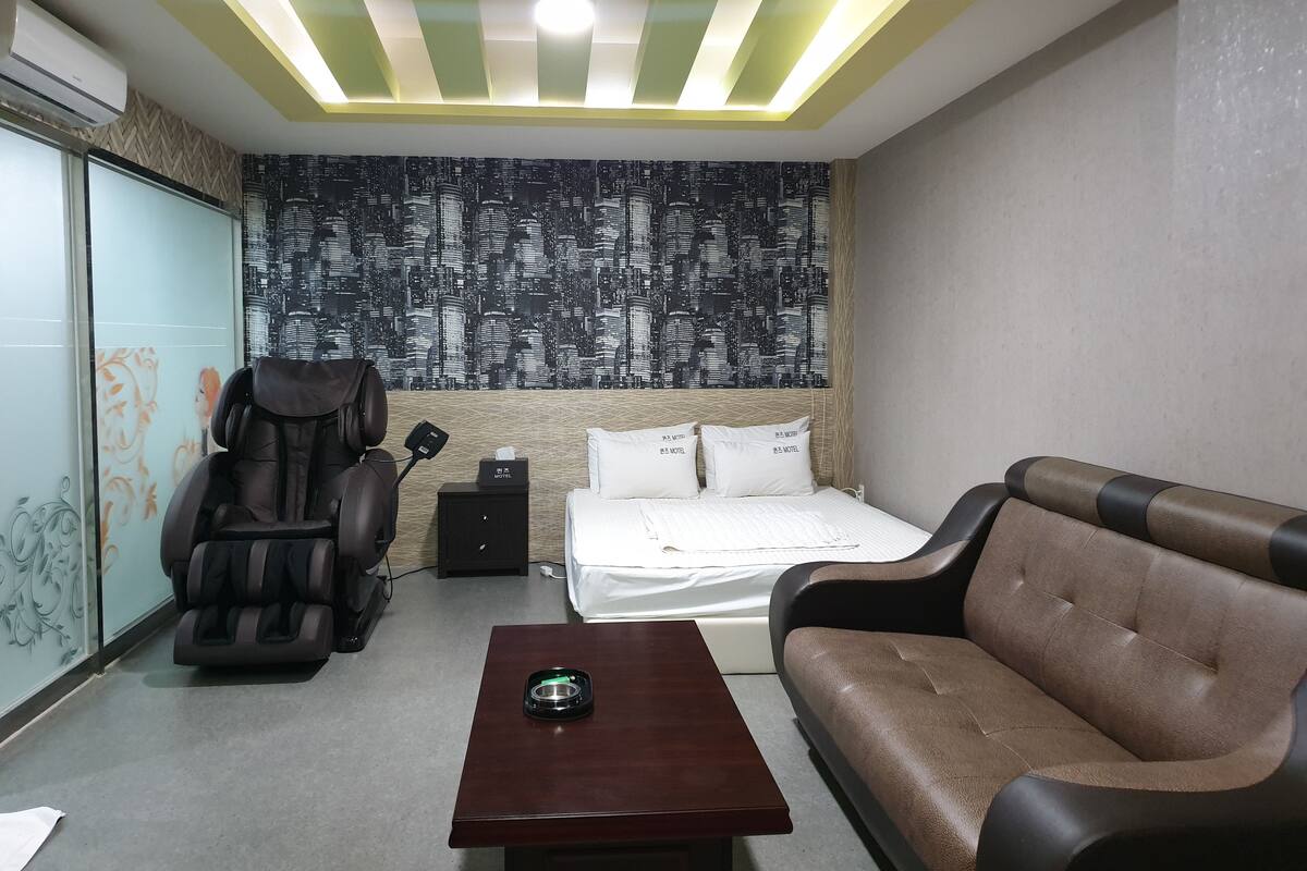 Quarto de hotel com cama, cadeira de massagem, mesa de massagem, mesinha e parede com um mural da paisagem urbana.