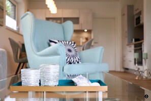 A modern living room with a light blue armchair, a glass coffee table with decorative items, and a view into the kitchen area. - Kapitänshaus 2 (Kellenhusen)