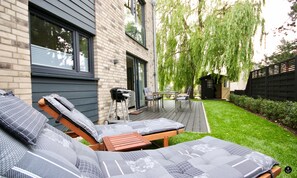 A modern outdoor patio with a wooden deck, a grill, and a seating area with cushioned loungers. - Kapitänshaus 2 (Kellenhusen)
