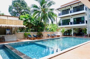 Piscina con sillones de descanso, rodeada de plantas tropicales y un edificio de varios pisos con balcones.