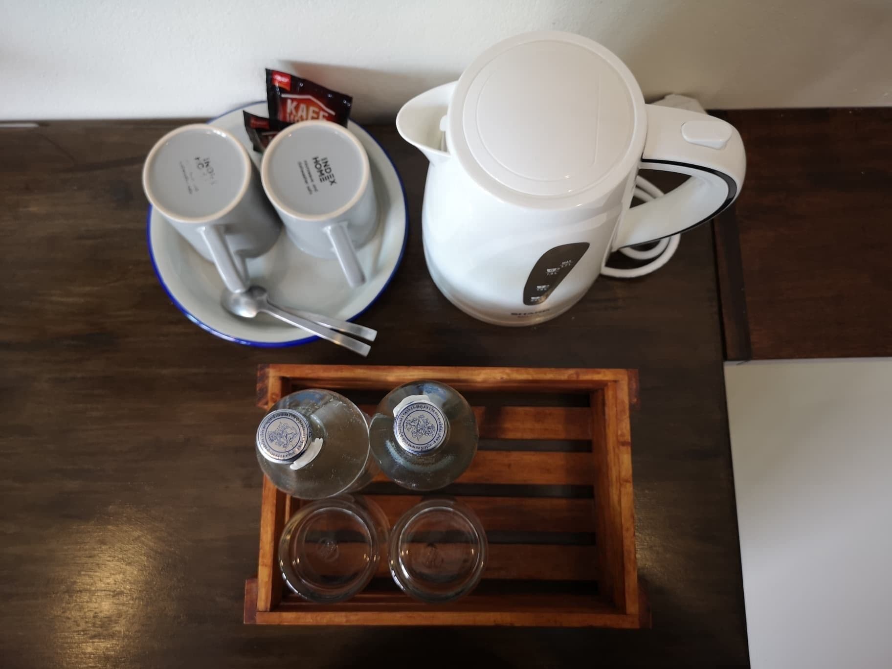 Eine Kaffeemaschine, zwei Tassen, ein Löffel und ein Tablett mit drei Gläsern auf einem Holztisch.