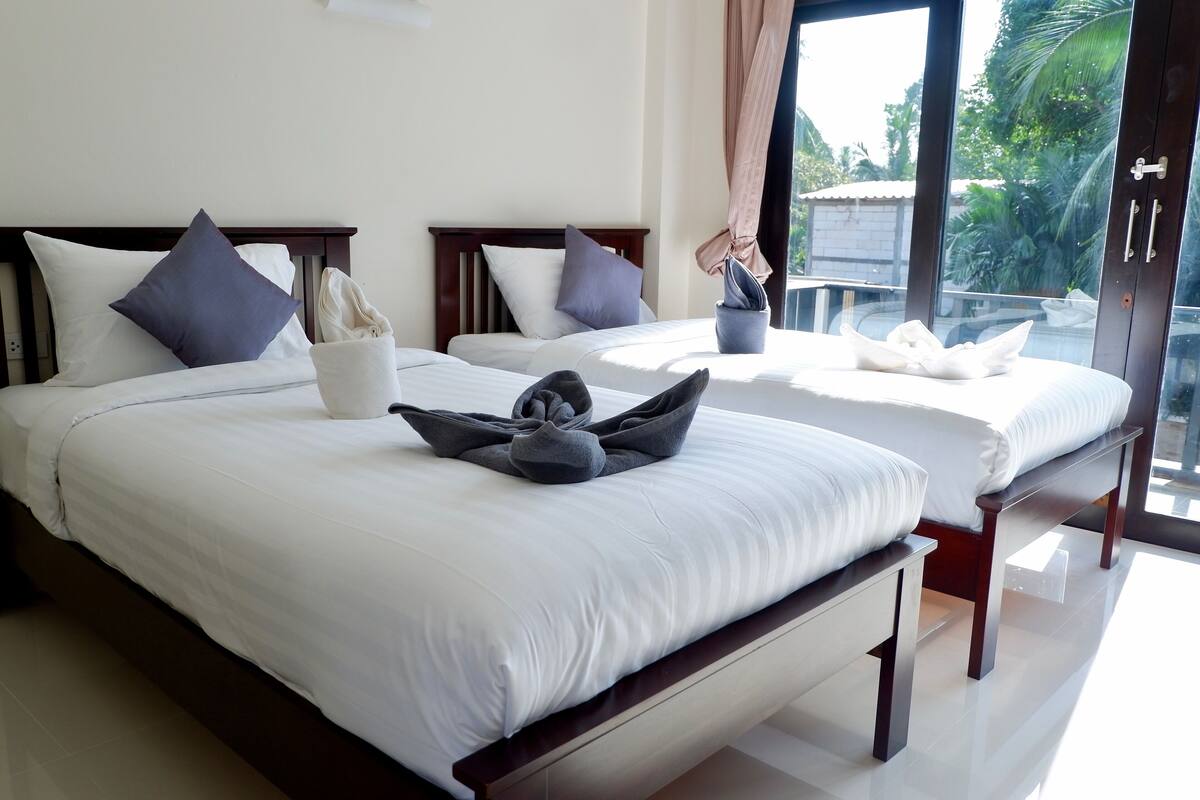 Une chambre d’hôtel avec deux lits, chacun doté d’une tête de lit en bois, de draps blancs et offrant une vue sur les palmiers à travers de grandes fenêtres.
