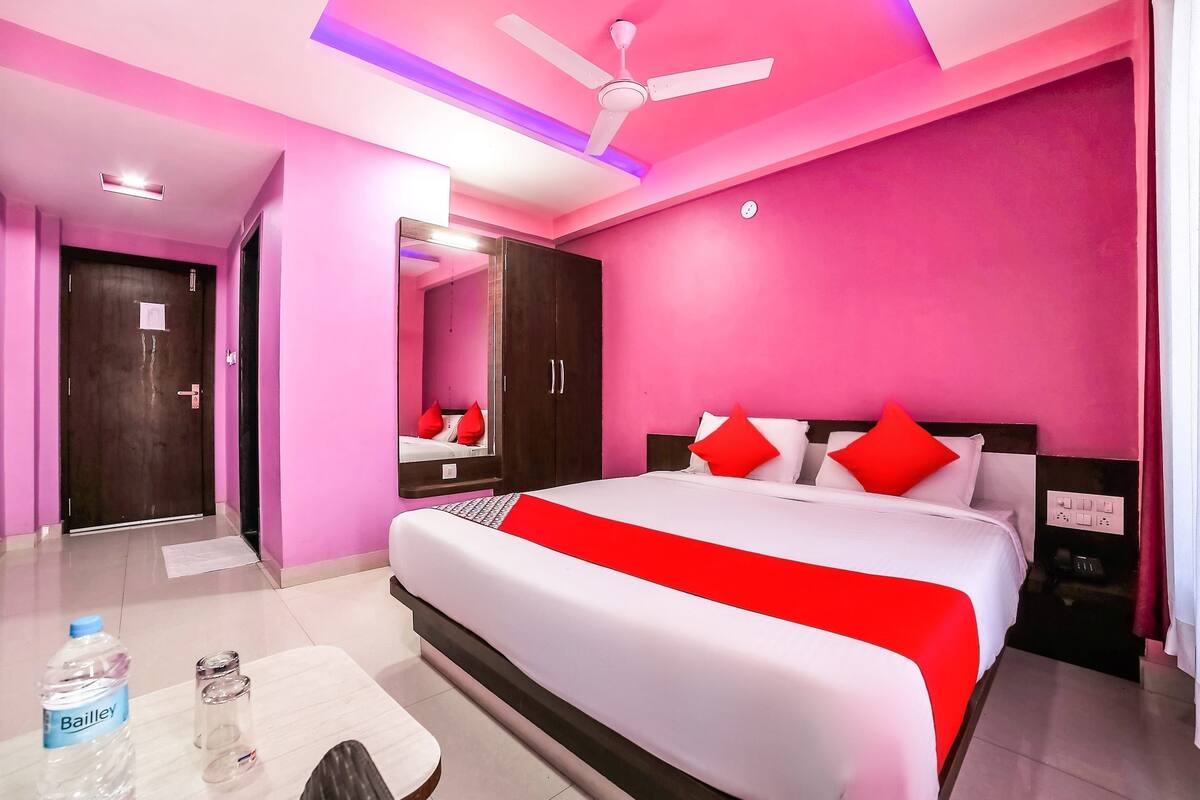 A hotel room with a large bed, a ceiling fan, a wardrobe, and a bedside table with a bottle and glasses.