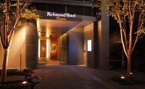 Vue de l’extérieur du Richmond Hotel Premier la nuit, avec une entrée moderne dotée de portes en verre, une enseigne lumineuse et un espace paysager agrémenté d’arbres.