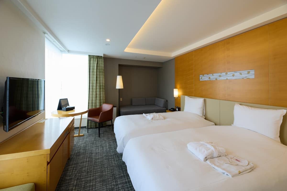 A hotel room with two beds, a desk with a TV, a chair, and a sofa.