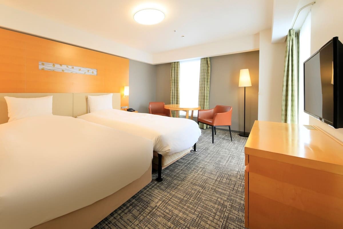 A hotel room with two beds, a desk, a chair, a TV, and a window with curtains.