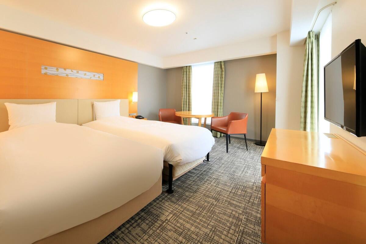 A hotel room with two beds, a desk, a chair, a TV, and a window with curtains.