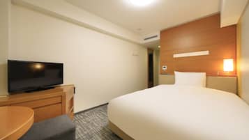 A hotel room with a bed, a TV on a wooden dresser, a desk with a chair, and a wall-mounted light.