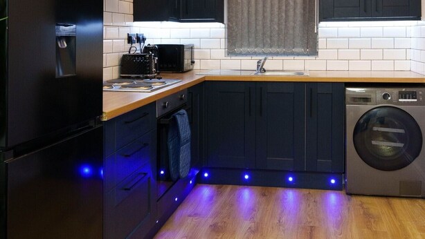 A modern kitchen with dark cabinets, a wooden countertop, a built-in oven, a washing machine, and under-cabinet lighting. - Spencer House (Leeds)