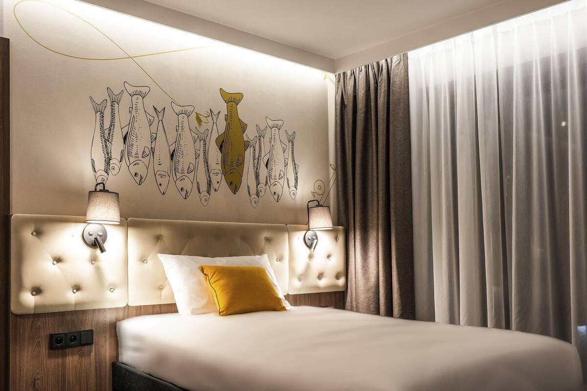 Ein Hotelzimmer mit einem gepolsterten Kopfteil, einem Bett mit weißen und senffarbenen Kissen, einem Wandbild von Fischen und einem großen Fenster mit Vorhängen.