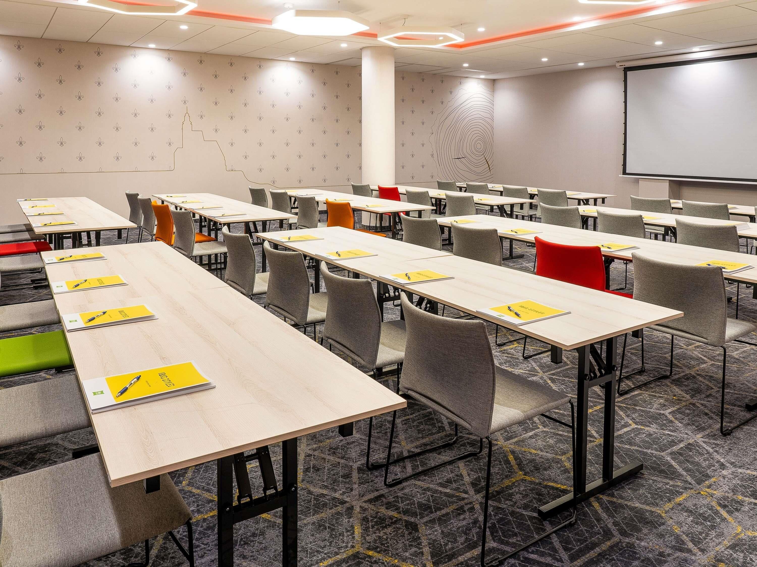 Une salle de conférence équipée de plusieurs tables et chaises, d’un écran de projection et d’un mur orné de motifs.