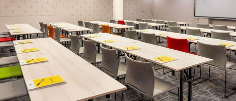 A conference room with multiple tables and chairs, a projector screen, and a patterned wall.