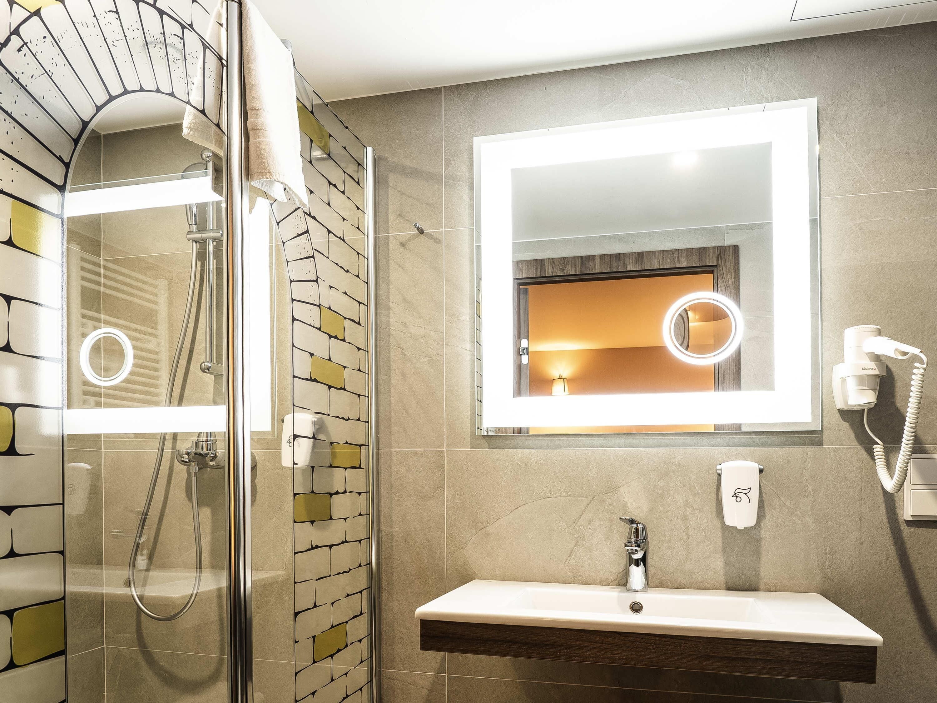 A bathroom with a glass shower enclosure, a sink with a mirror, and a hairdryer mounted on the wall.