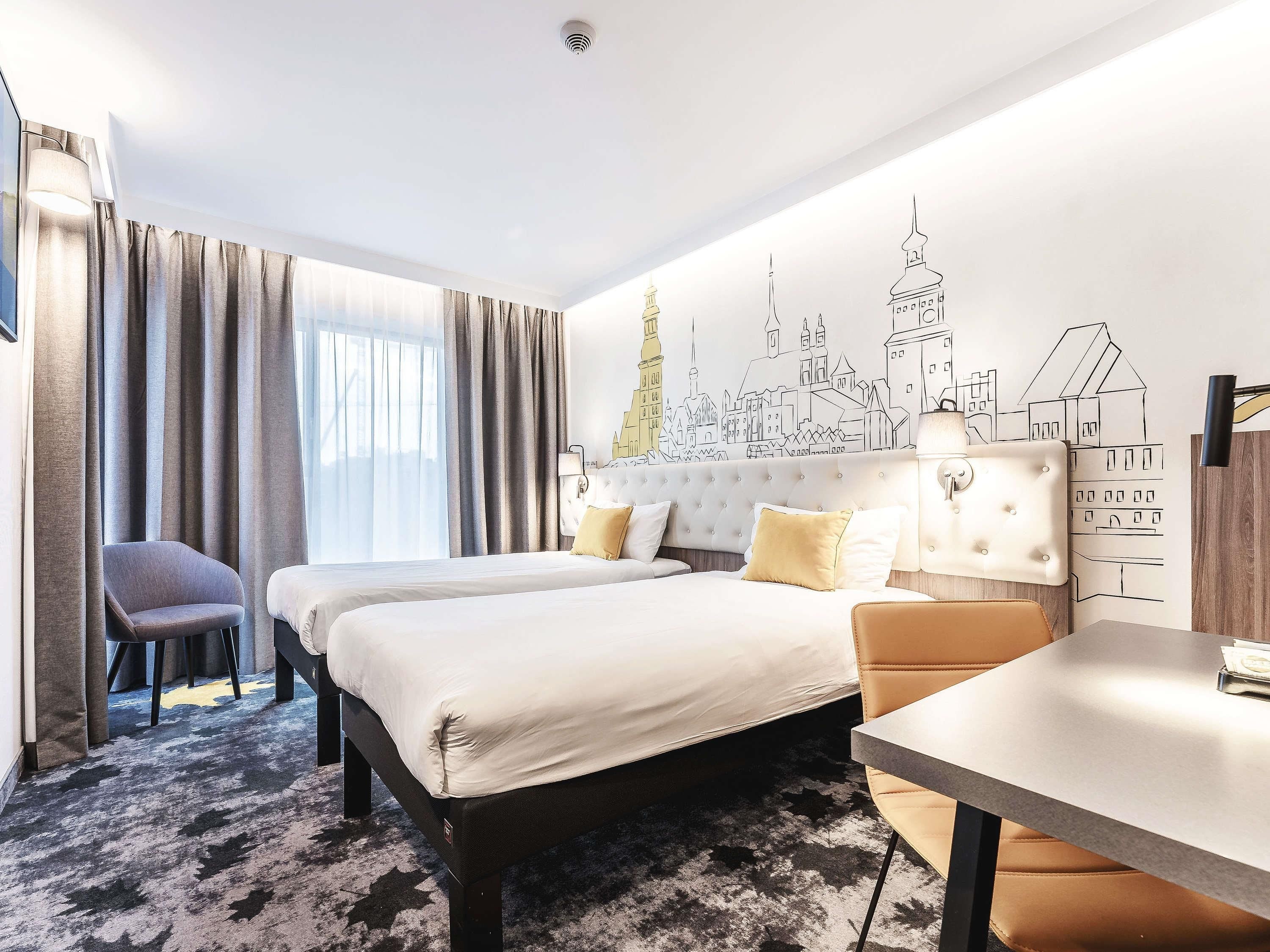 A hotel room with two beds, a desk, a chair, and a large window with curtains.