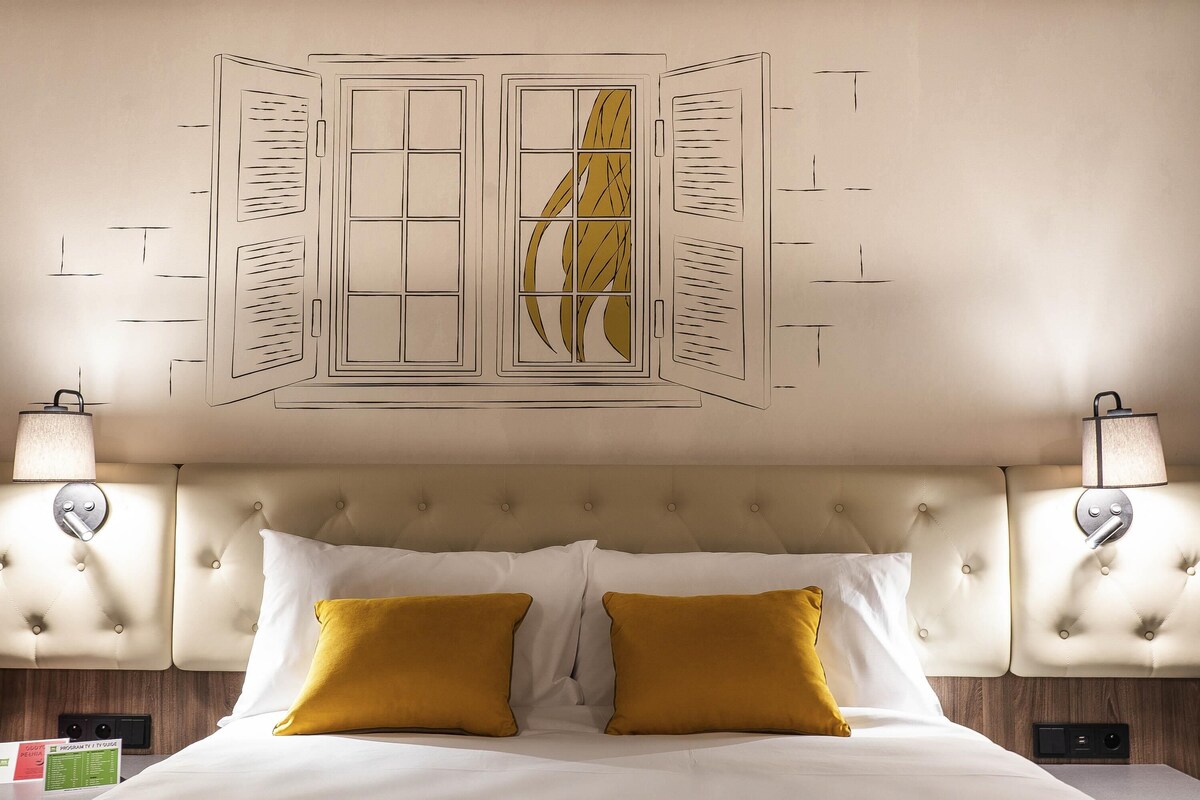 A hotel room with a tufted headboard, two bedside lamps, a bed with white and mustard pillows, and a wall art piece above the bed.