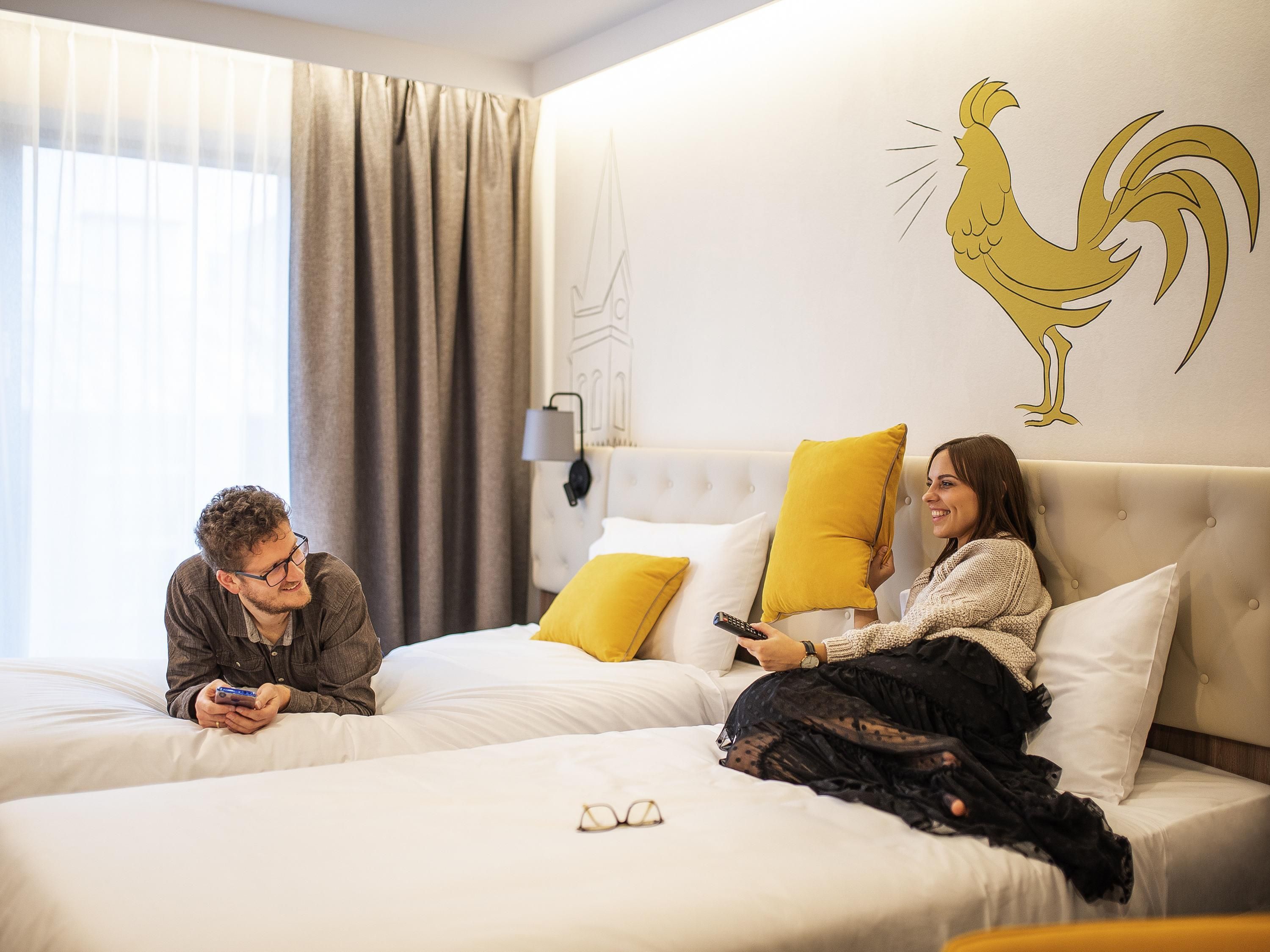 A hotel room with two beds, a large window with curtains, a wall-mounted lamp, and a rooster decal.