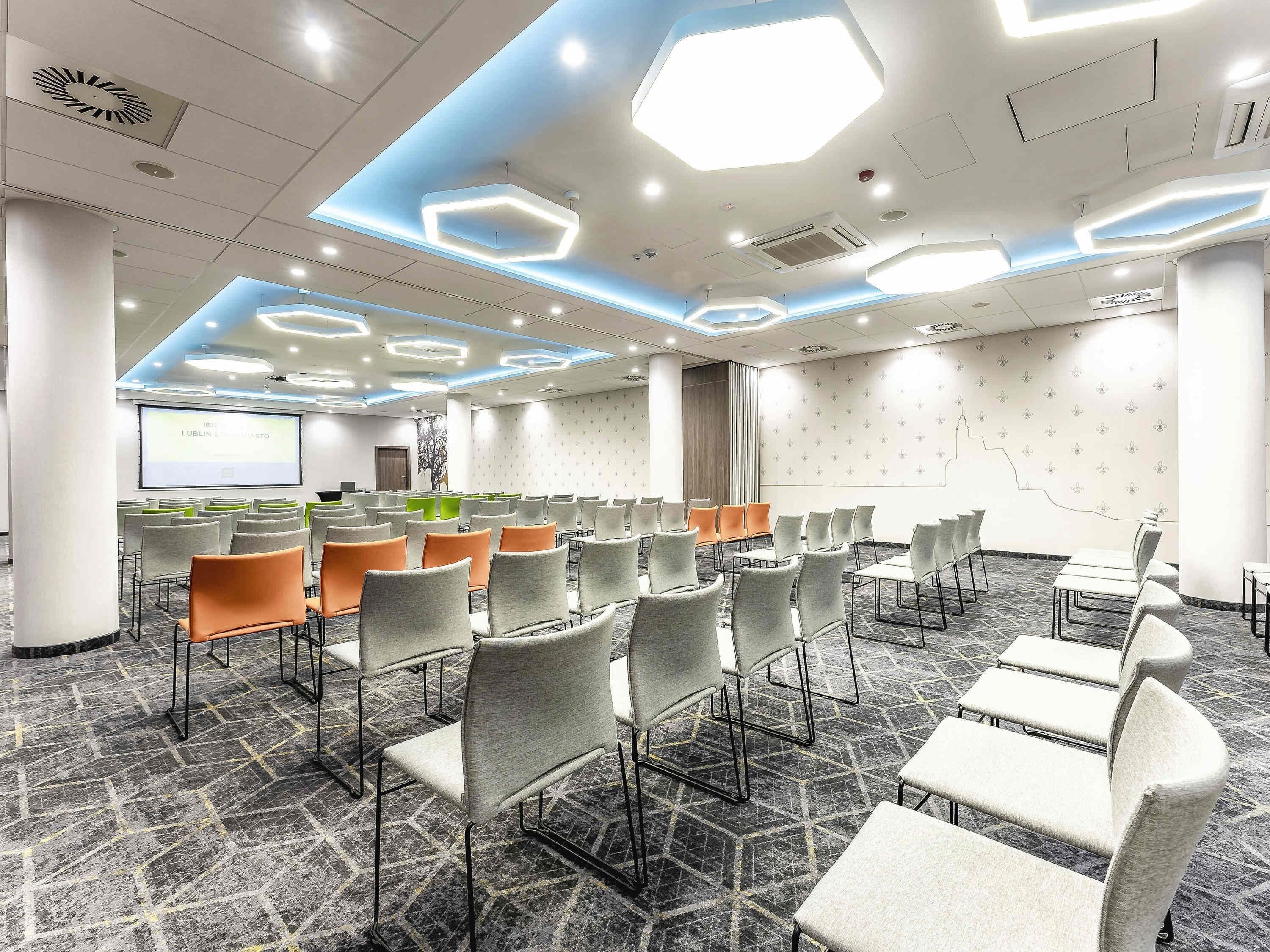 A conference room with a modern design, featuring a large screen, multiple rows of chairs, and a patterned carpet.