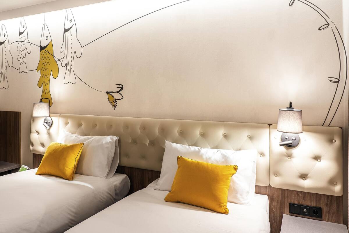 Ein Hotelzimmer mit einer gebügelten Kopfstütze, zwei Betten mit weißen und gelben Kissen sowie einer Wandmalerei und einer Nachttischlampe.