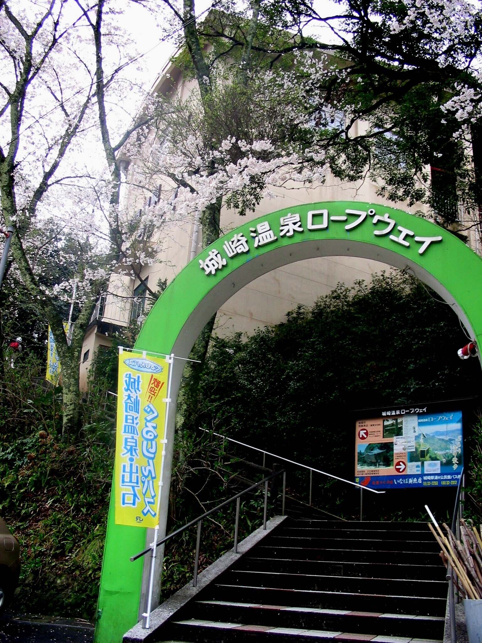 A green archway with Japanese text, leading to a staircase surrounded by trees and greenery.