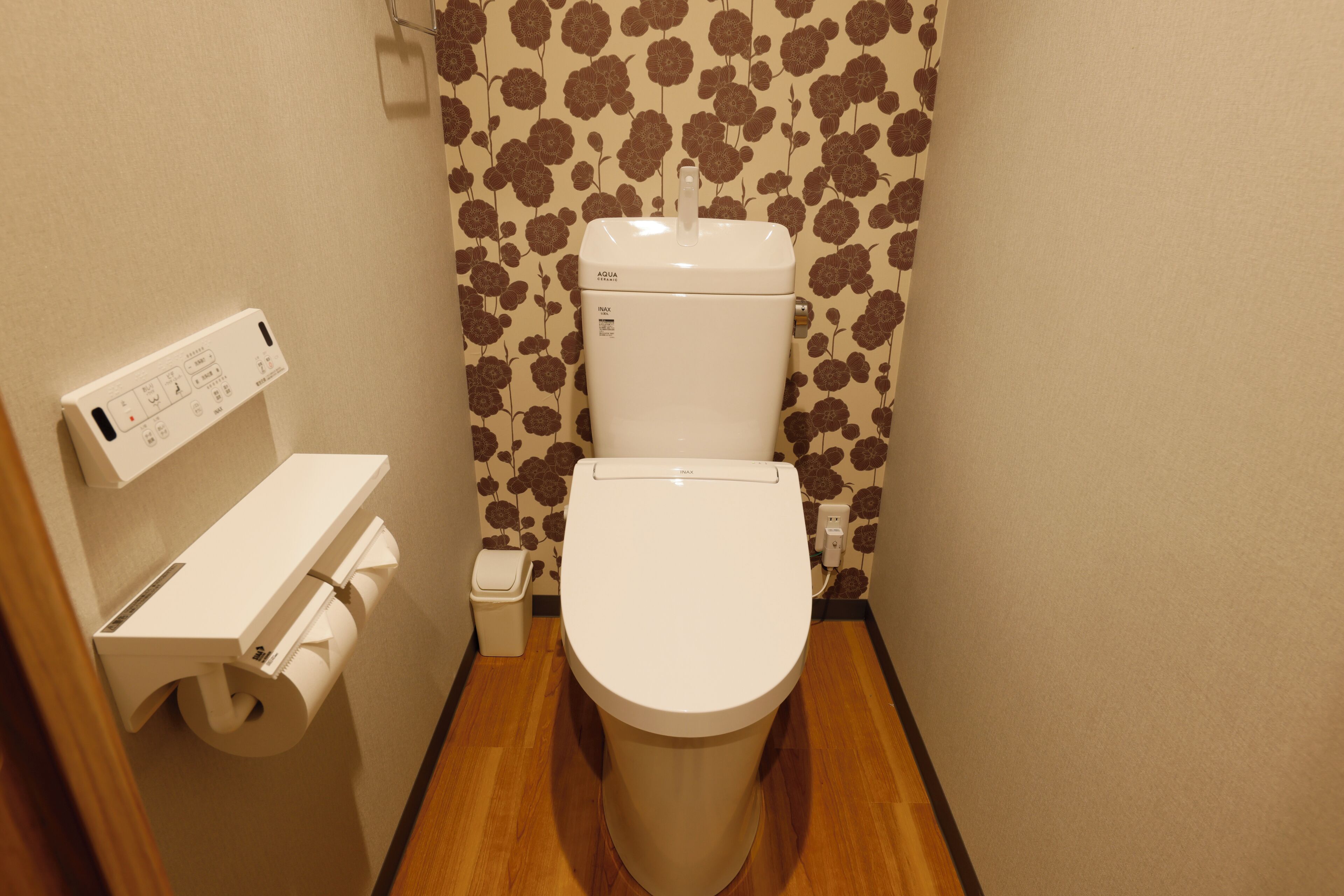 A small bathroom with a toilet, a paper towel dispenser, and a wall-mounted electrical outlet.