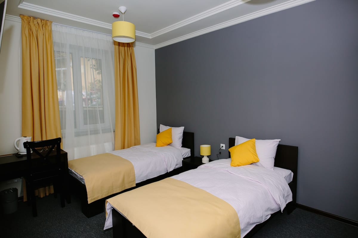 A hotel room with two single beds, a desk, a chair, a lamp, and a window with curtains.