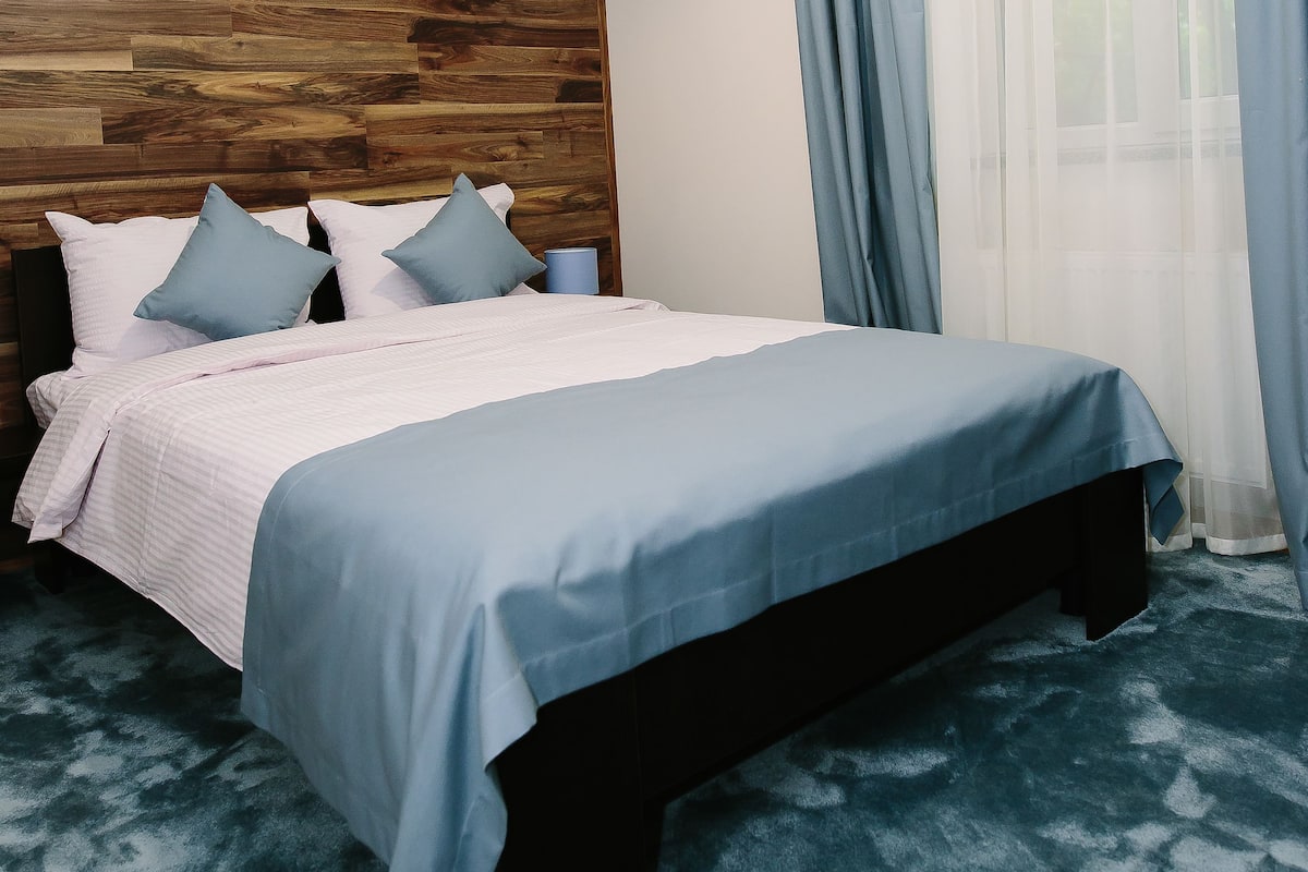 A bedroom with a wooden headboard, a bed with blue bedding, and a large window with blue curtains.