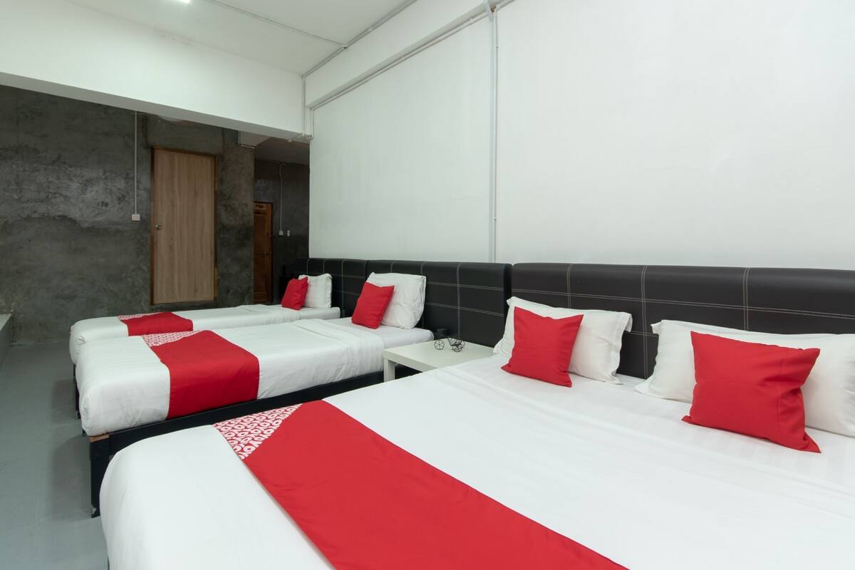 Une chambre d’hôtel avec deux lits, chacun agrémenté de draps blancs et de touches de couleur rouge. On y trouve une porte en bois et un mur blanc avec une fenêtre.