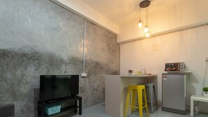A compact living space with a flat-screen TV on a stand, a small stool, a refrigerator, and a kitchenette area with a toaster and a plant. - OYO 43993 Madsuite (Kota Kinabalu)