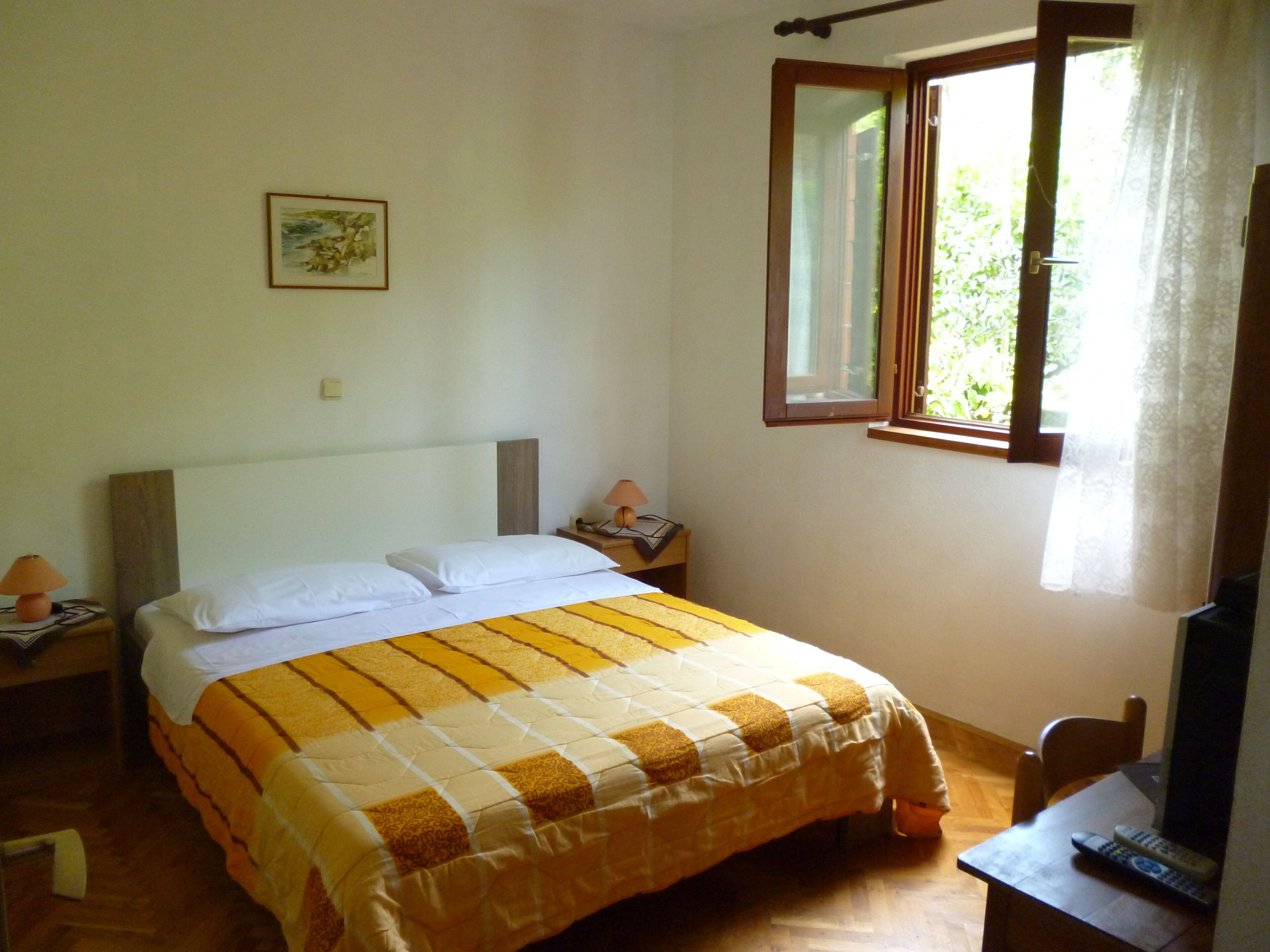 A bedroom with a bed, a nightstand, a lamp, a window with curtains, and a painting on the wall.