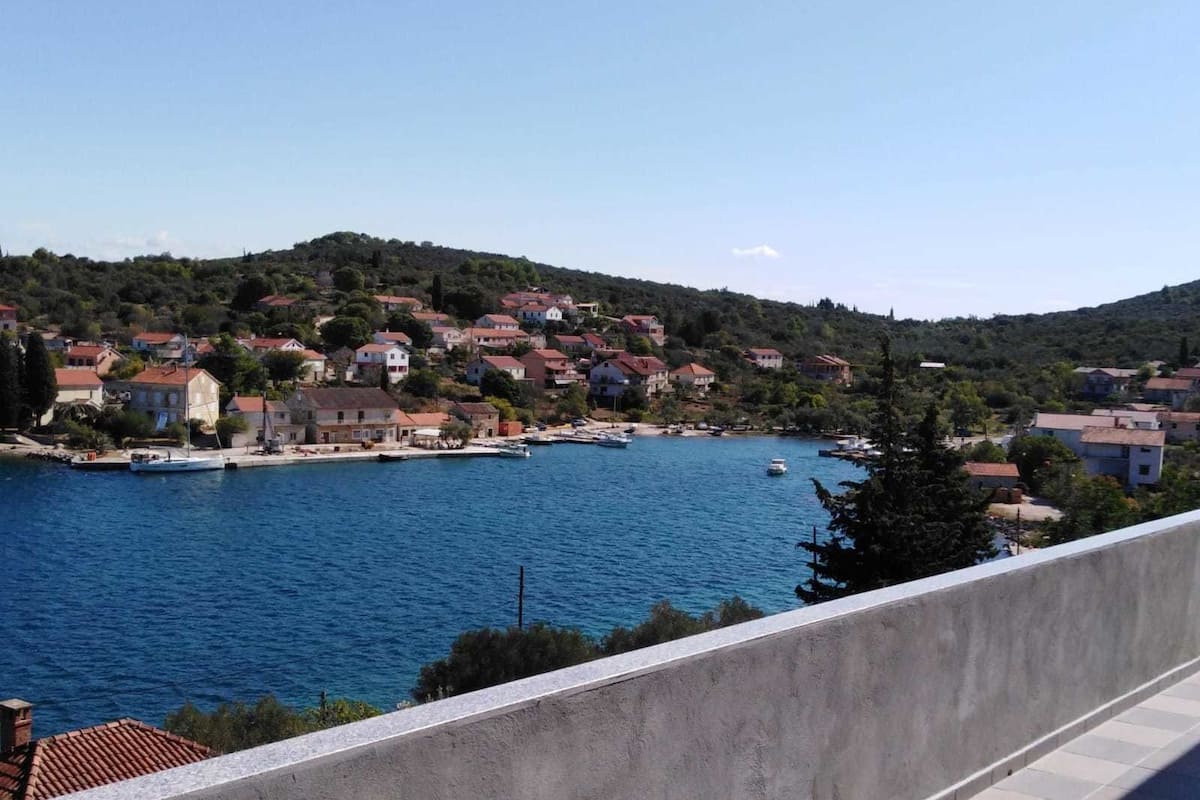 A coastal town with houses along the waterfront, boats docked, and a hilly backdrop.