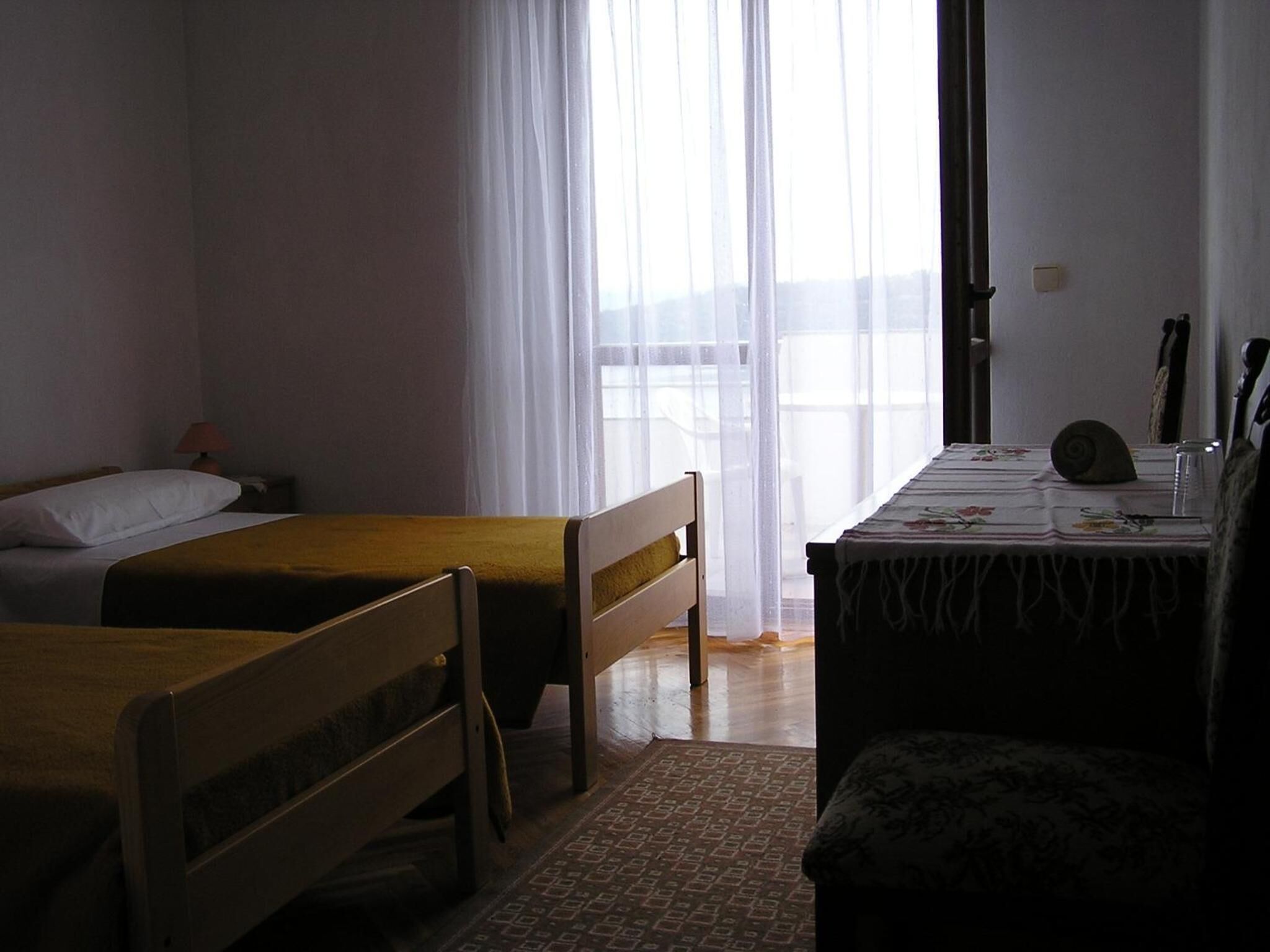 Ein Zimmer mit zwei Einzelbetten, einem kleinen Tisch mit Tischdecke, einem Stuhl und einem Fenster mit durchsichtigen Vorhängen.