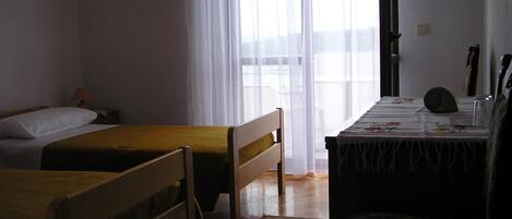 Quarto com duas camas de solteiro, uma mesinha com toalha, uma cadeira e uma janela com cortinas leves.